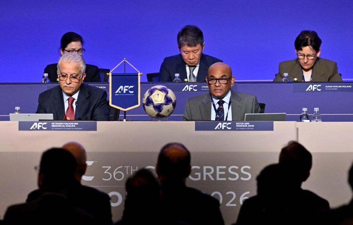 Panel of officials at a blue AFC conference stage, a soccer ball sits between two men at the front table with audience in foreground
