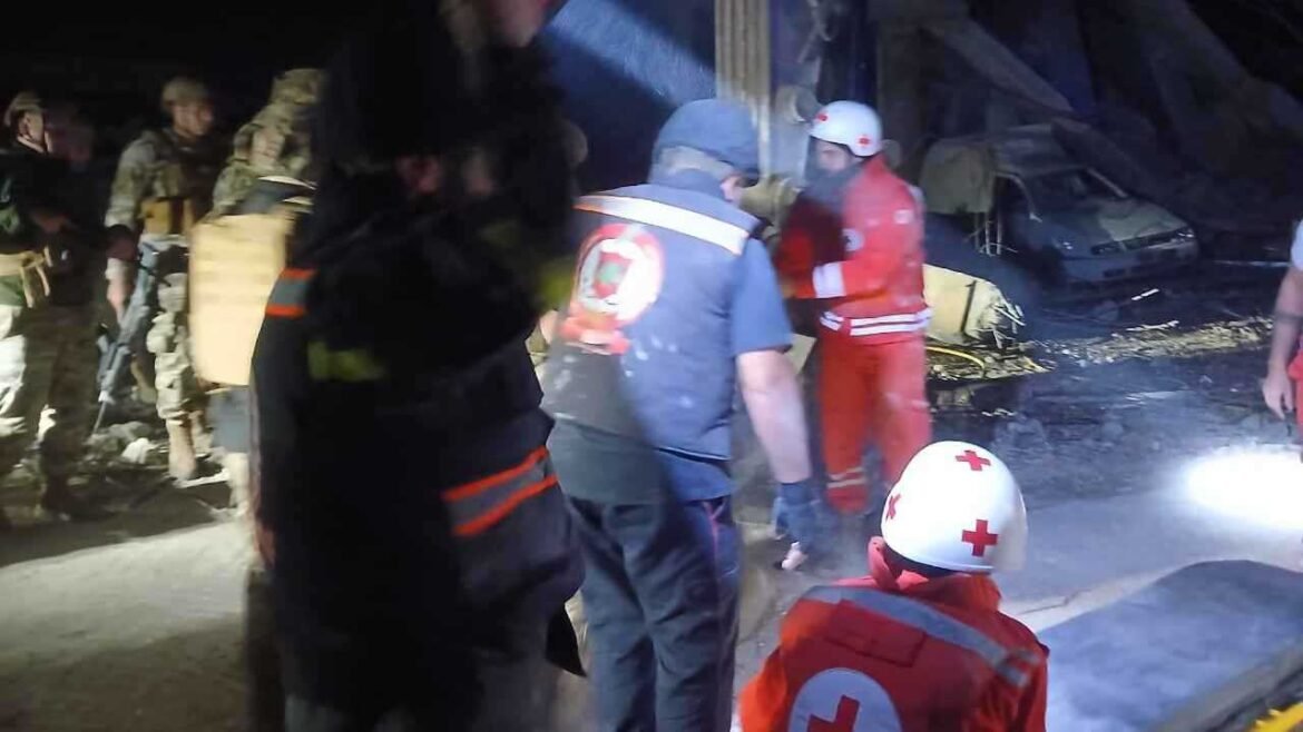 Nighttime disaster scene with emergency responders in red cross gear and helmets assisting amid rubble and a damaged vehicle.