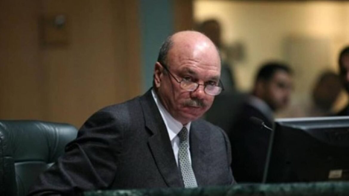 Man in a dark suit and patterned tie sits at a conference table in a boardroom, looking over his glasses toward the camera.