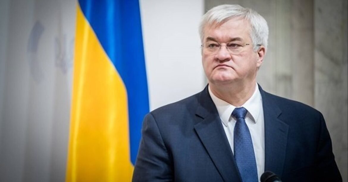 Man in a dark suit and blue tie speaks at a podium, Ukrainian flag in the background.