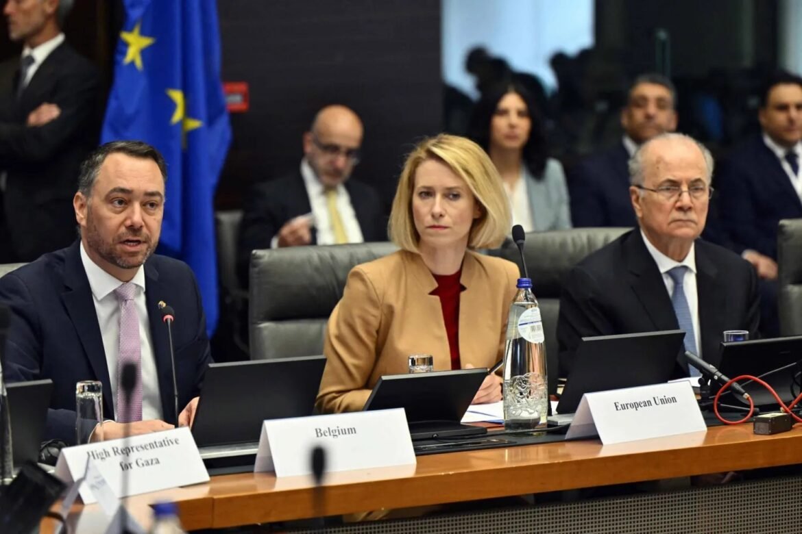 Three officials sit at a formal conference table with microphones, EU flag in the background and country and EU nameplates visible (Belgium, European Union).