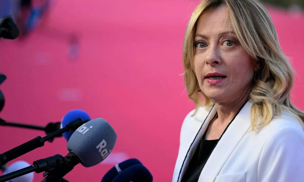 Close-up of a blonde woman in a white blazer speaking at a press conference, with microphones in the foreground and a pink background