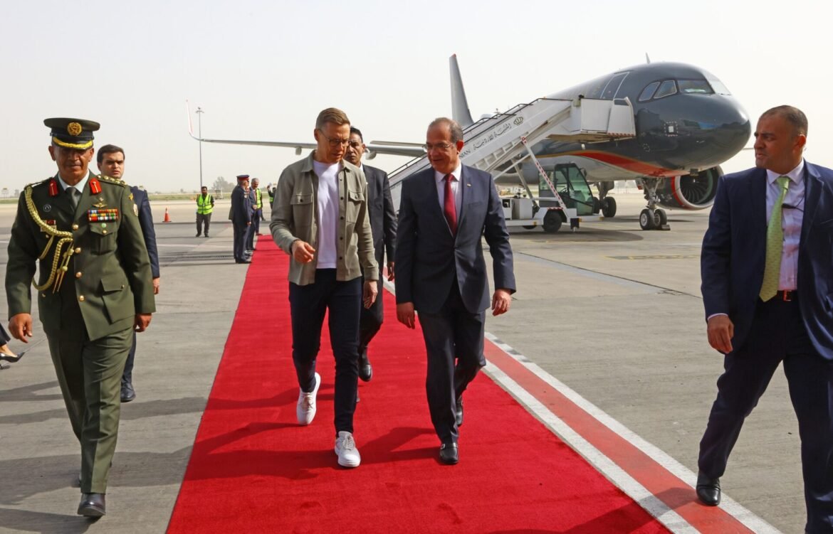 Group of officials walking on a red carpet at an airport, a military officer in green uniform leading alongside suited men and a passenger jet in the background.