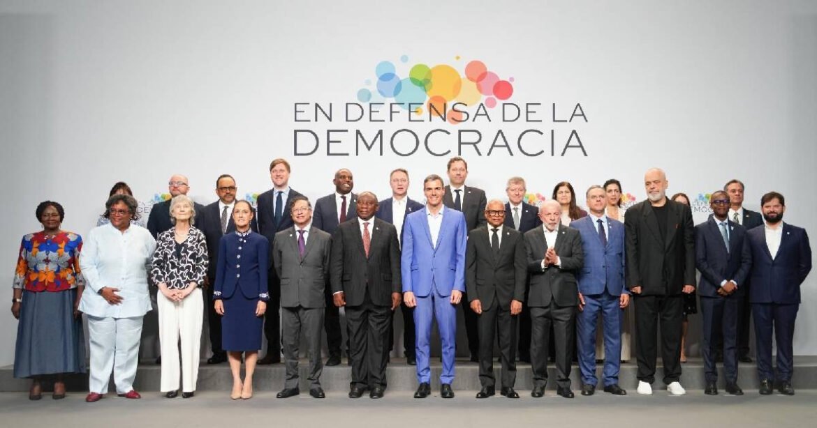 Diverse group of officials in formal attire posing on a stage in front of a 'En defensa de la democracia' banner, conveying a democracy event.