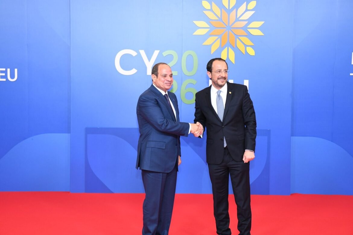 Two men in dark suits shake hands on a red carpet against a blue backdrop with a yellow geometric emblem and text behind them.
