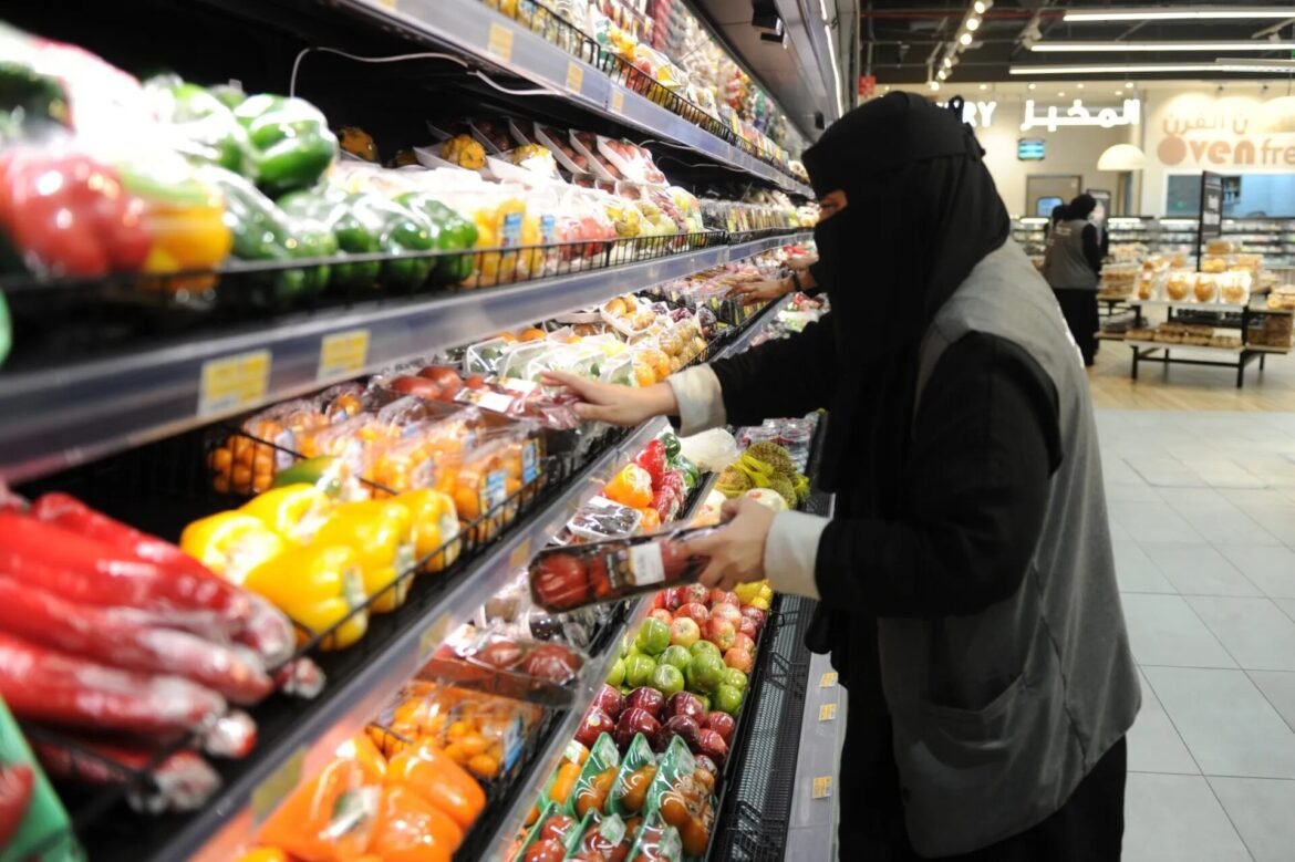 Woman wearing a black hijab and coat selecting packaged fruit in a supermarket produce aisle.