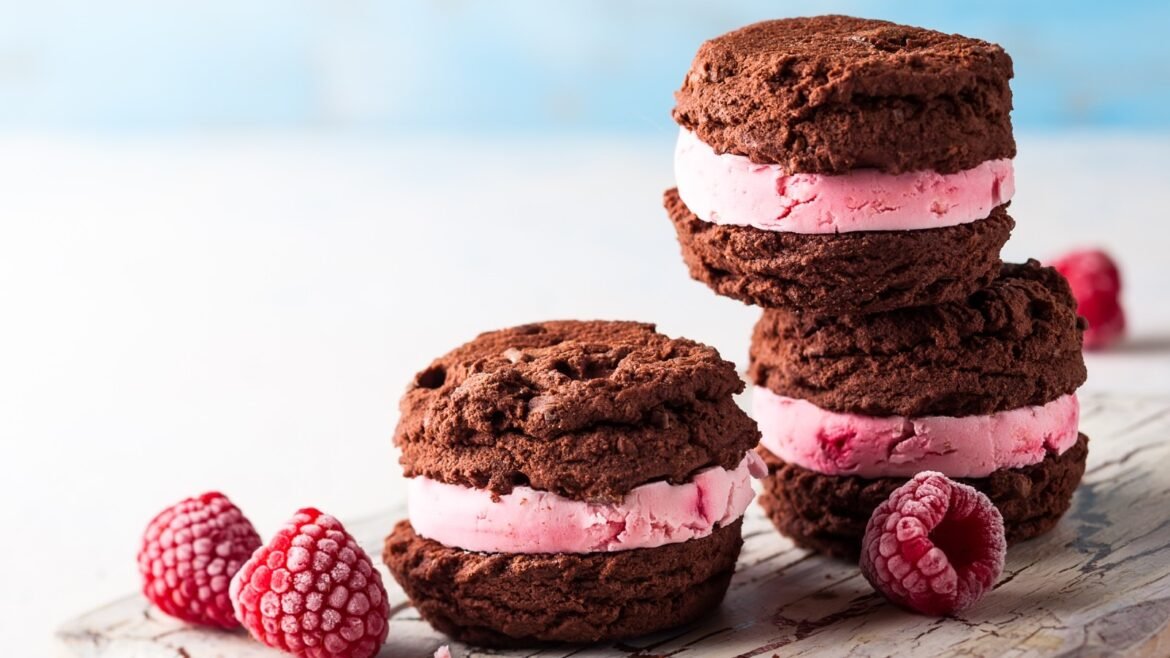 Stack of chocolate cookies with pink cream filling stacked on a wooden board, with fresh raspberries nearby.