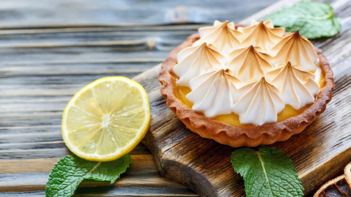 Lemon tart with piped toasted meringue on a wooden board, beside a lemon half and mint leaves.