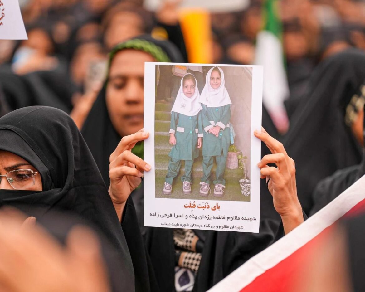 A woman in a black hijab holds a poster showing two young girls in green school uniforms and white headscarves at a crowded gathering, with Persian text beneath the image.