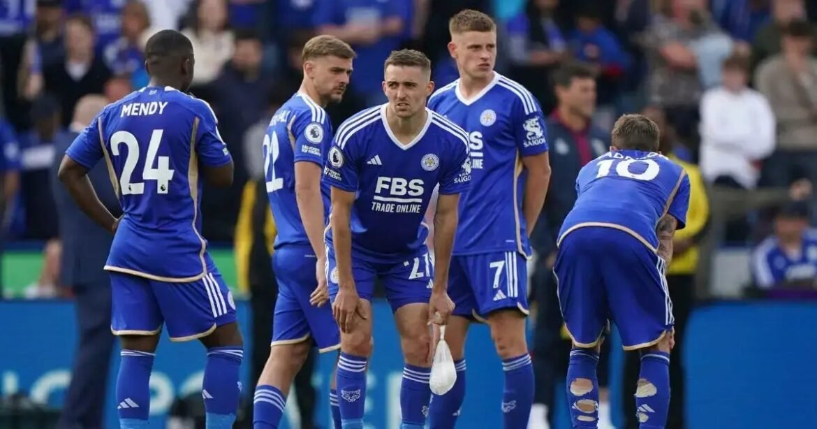 Group of football players in blue kits on the pitch during a match, looking focused and halted in a moment of play.