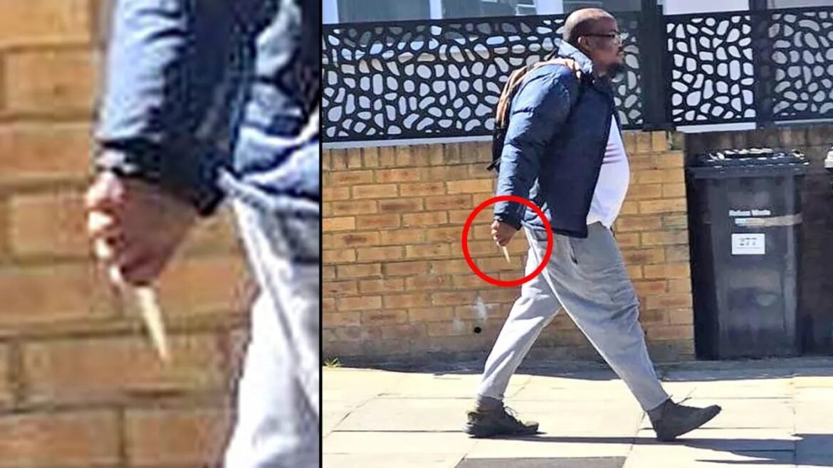 Man in a blue jacket and gray pants walks on a sidewalk with a backpack, holding a slim object in his right hand against a brick wall and a trash bin in the background.