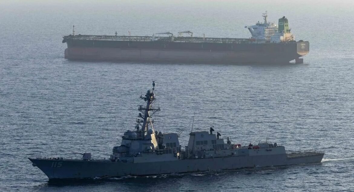 Military gray destroyer sailing in open water with a large cargo ship visible on the horizon.