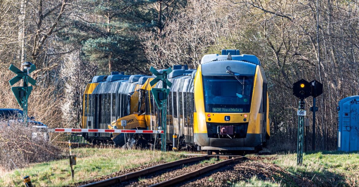 Yellow passenger train derailed on tracks near a crossing, with damaged cars and barriers in a wooded area.