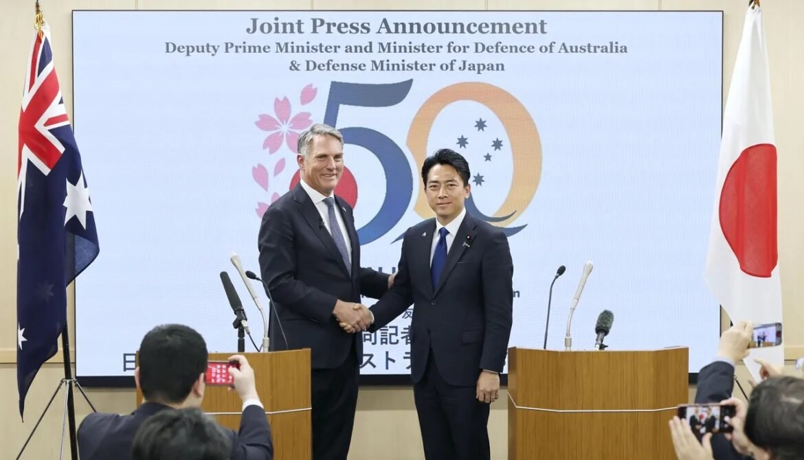 Two men in dark suits shake hands at a joint press conference, with Australian and Japanese flags and a large screen showing a 50th-anniversary design behind them.