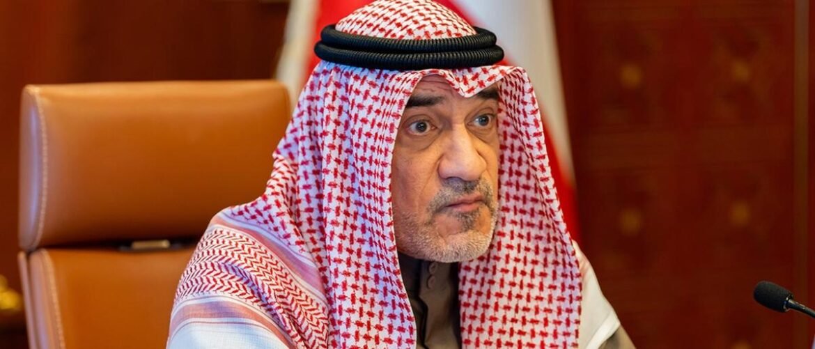 Middle-aged man in a red-and-white keffiyeh and dark thawb sitting at a conference table, looking thoughtful, with a microphone nearby and warm wood panel background.