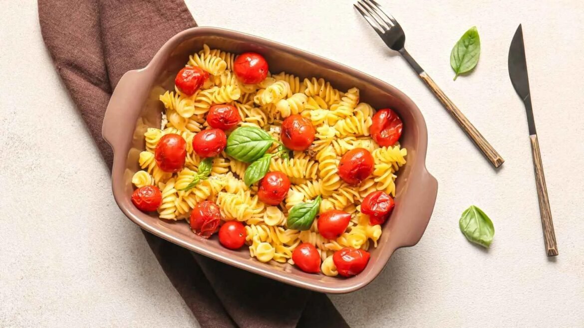 Brown baking dish filled with fusilli pasta, cherry tomatoes, and fresh basil on a light countertop.