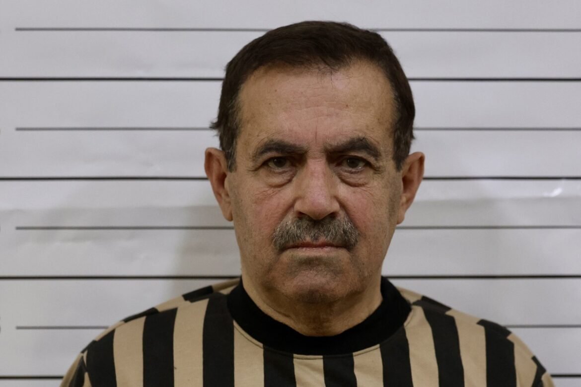 Mugshot of a middle-aged man with a mustache in a beige and black striped prison uniform, standing against a height measurement backdropok behind him.