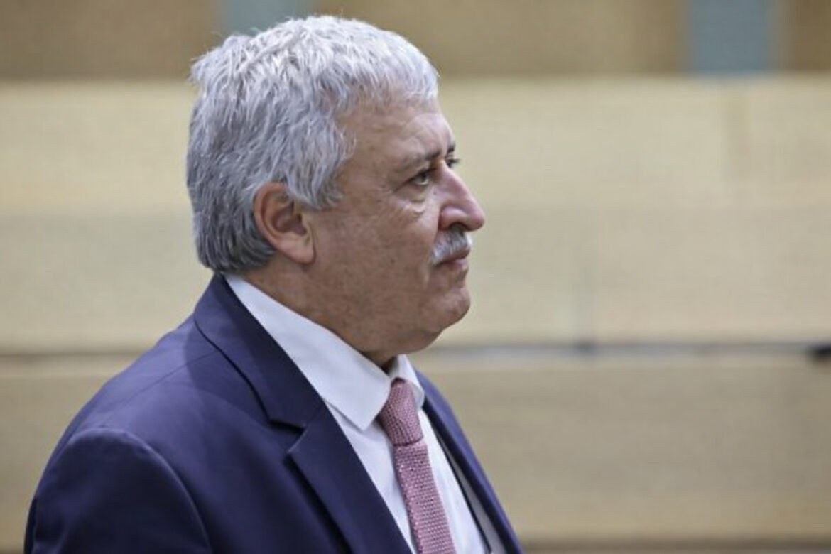 Side profile of an older man with gray hair in a navy suit and pink tie, outdoors.