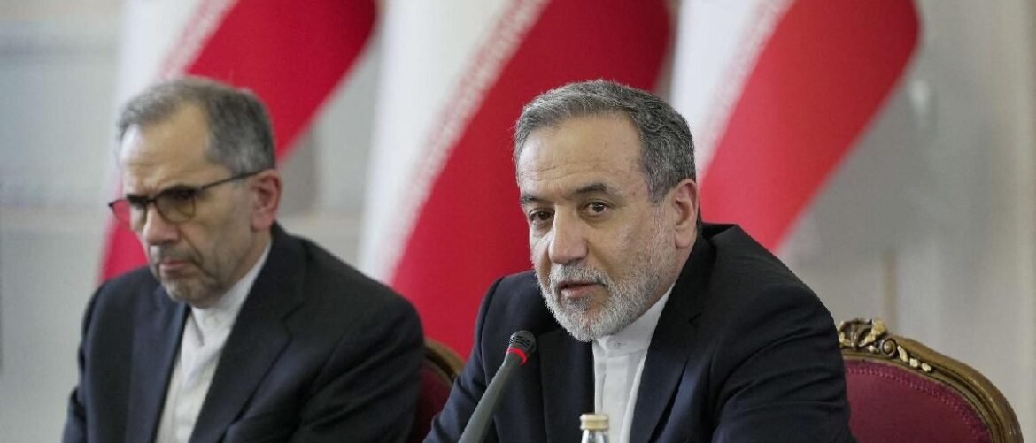 Two men in suits at a formal press conference, one speaking into a microphone with red-and-white flags in the background.