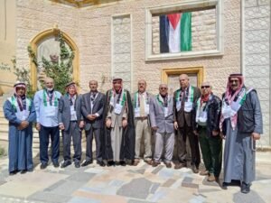 Group of Men in Suits and Traditional Robes Posing for a Formal Photo in Front of a Stone Building with a Palestinian Flag Display Above a Doorway