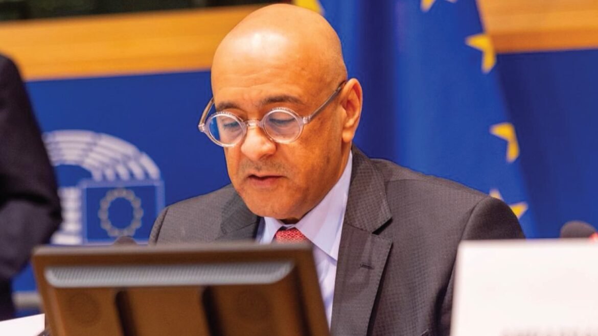 Bald man with round glasses in a gray suit and red tie, reading at a desk in a formal meeting room with blue EU backdrop behind him.