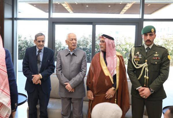 Four men stand indoors in a modern lobby; two in suits, one in traditional Arab attire, and a military officer in a green beret. Behind them are large glass doors.