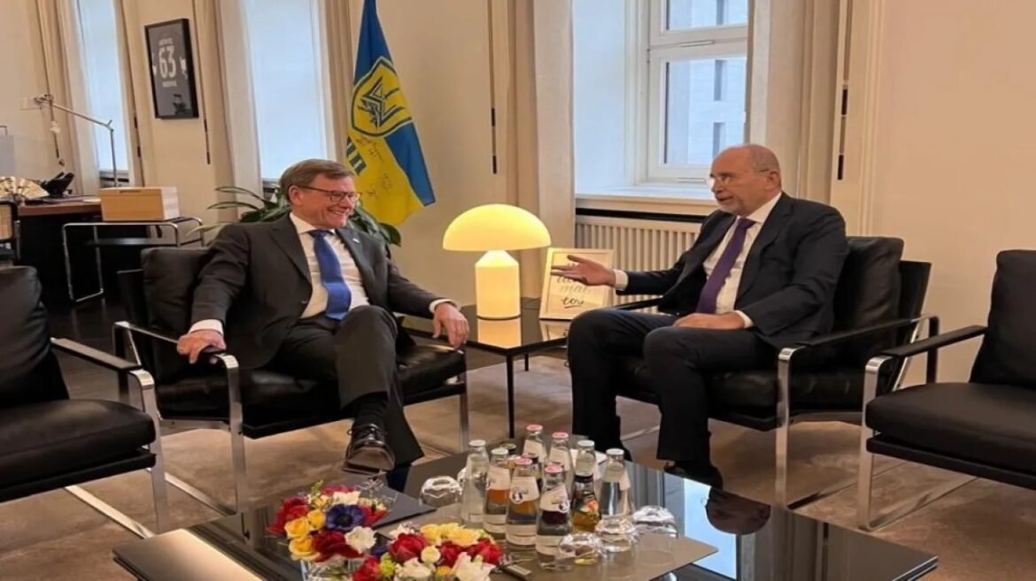 Two suited men converse in a bright office, Ukrainian flag in the background, with a glass coffee table holding bottled drinks and flowers.