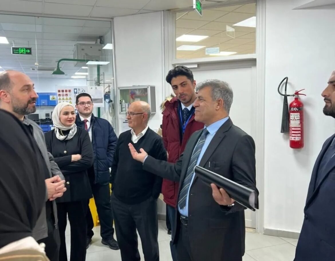 Group of professionals in a lab discussing; a man in a suit gestures while holding a rolled document, as others listen attentively near a fire extinguisher on the wall