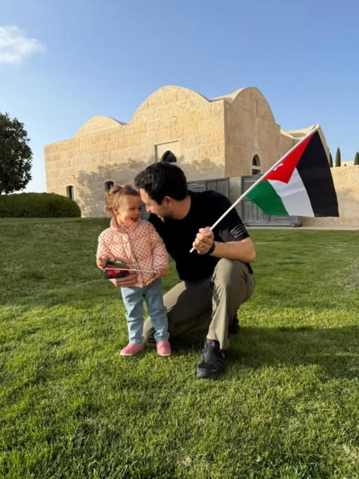 Man kneels on green grass with a little girl, both smiling, holding a Palestinian flag near a beige stone building.