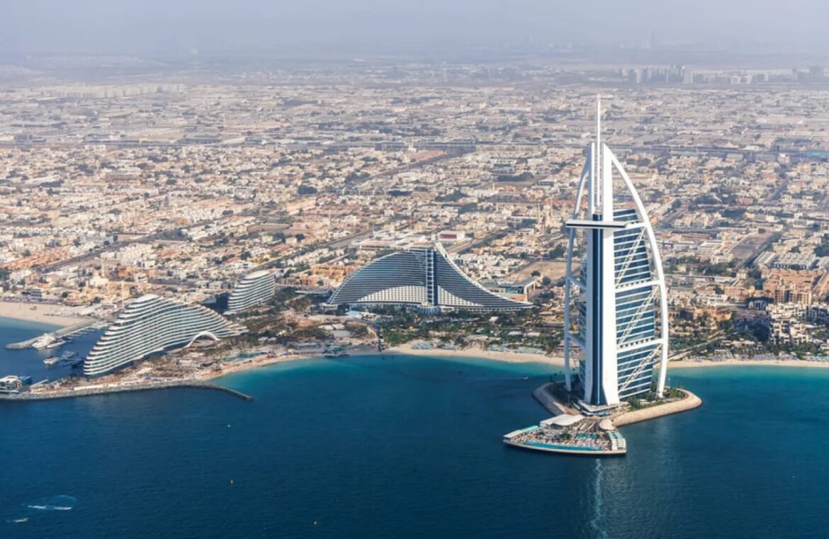 Aerial view of Dubai coastline showing the Burj Al Arab on its artificial island beside curved white hotels along the shore.