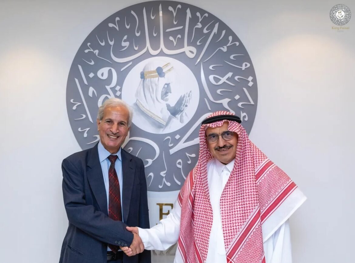 Two men in a suit and traditional Saudi attire shake hands in front of a circular Arabic calligraphy emblem on the wall.
