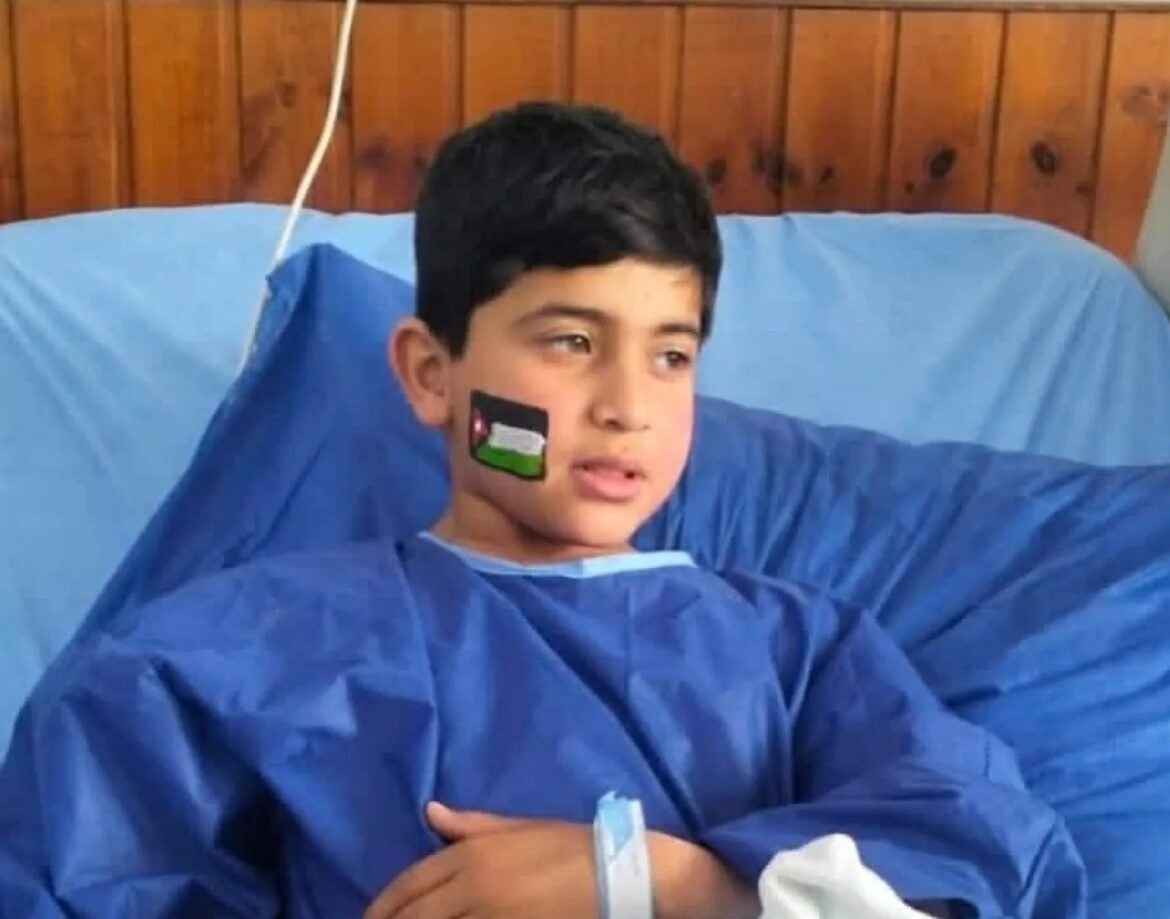 Boy in a blue hospital gown resting in bed with a flag sticker on his cheek.