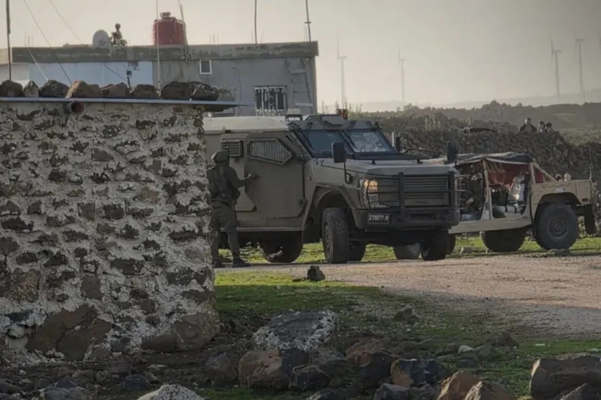 Soldier in tactical gear standing beside an armored vehicle near a rough stone wall in a rocky field at a distance.