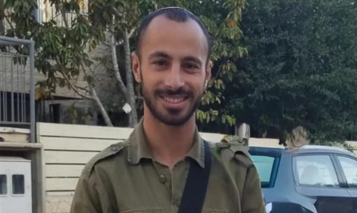Smiling man in an olive green shirt with a crossbody strap, standing outdoors with trees and a blue car behind him.
