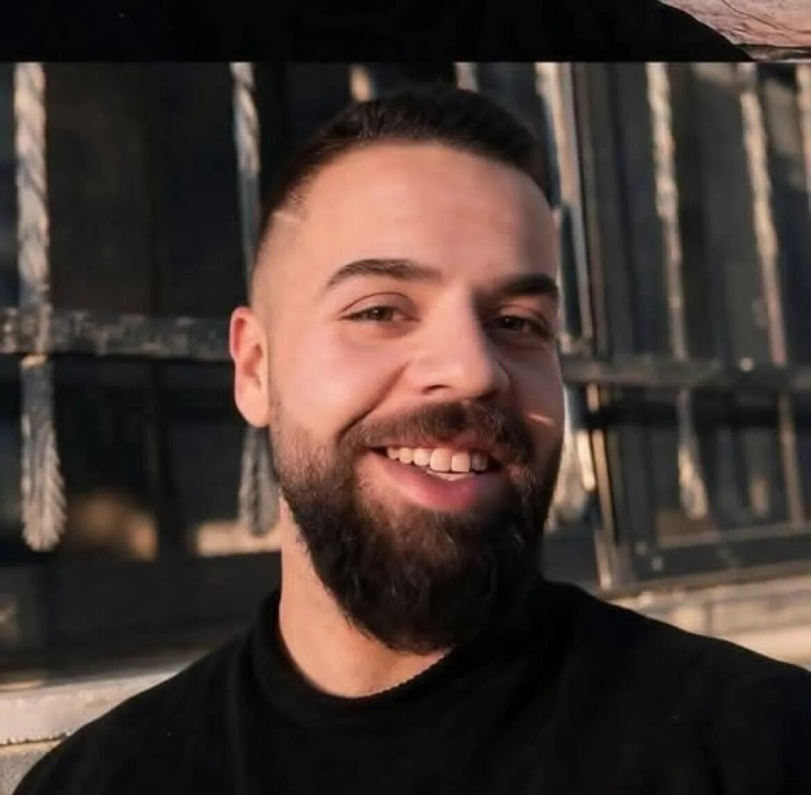 Smiling man with a dark beard and short hair, wearing a black shirt, outdoors with wooden background behind him.