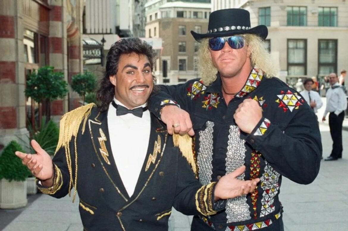 Two Elvis-style performers posing together on a city street, one in a black fringed jacket and bow tie, the other in a studded shirt with a cowboy hat and sunglasses.