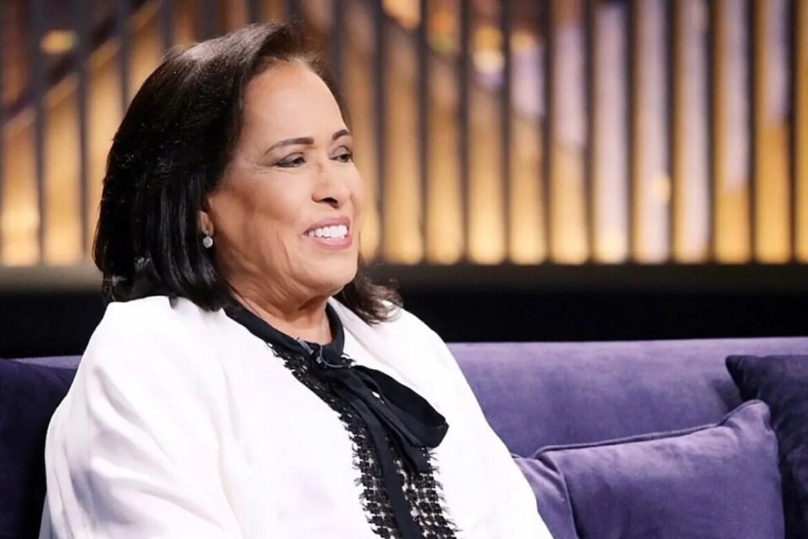 Smiling woman with dark hair in a white blazer seated on a purple couch during an interview setting.