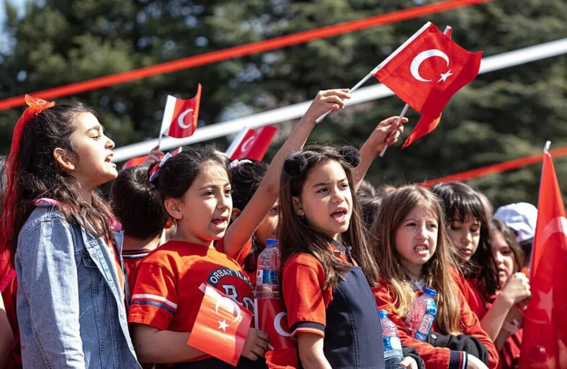 Group of children cheering at a Turkish flag parade, many waving small Turkish flags and holding water bottles.