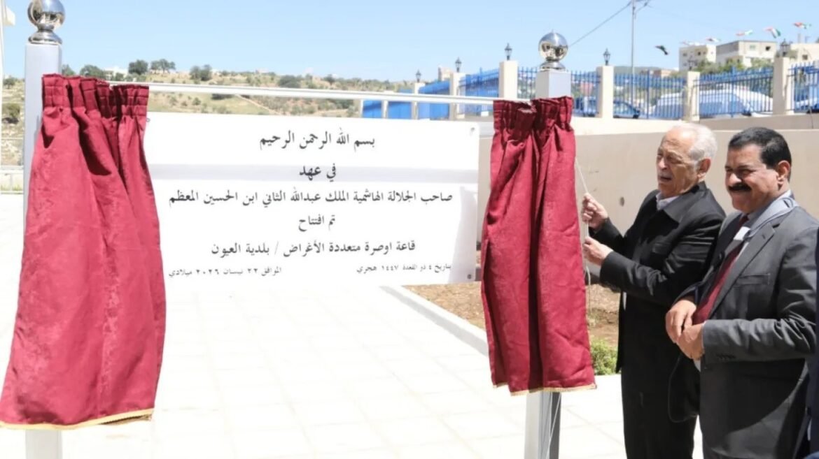 Two men in suits unveil a white sign mounted on a metal frame during a ceremony, maroon curtains pulled aside on both sides.