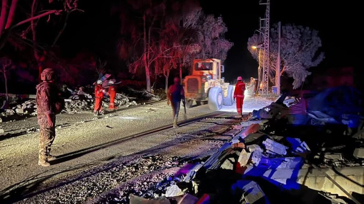 Nighttime accident scene with an orange excavator, debris along a damaged road and railway, and workers in reflective gear inspecting the site.” (informative)