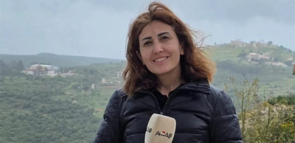 Female journalist outdoors, smiling, holding a beige microphone with Arabic logo against a hilly landscape.