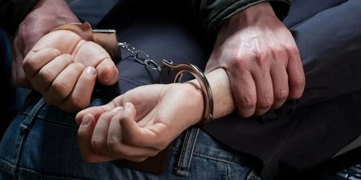 Person with hands cuffed behind their back being held by another person’s hands in a close-up.