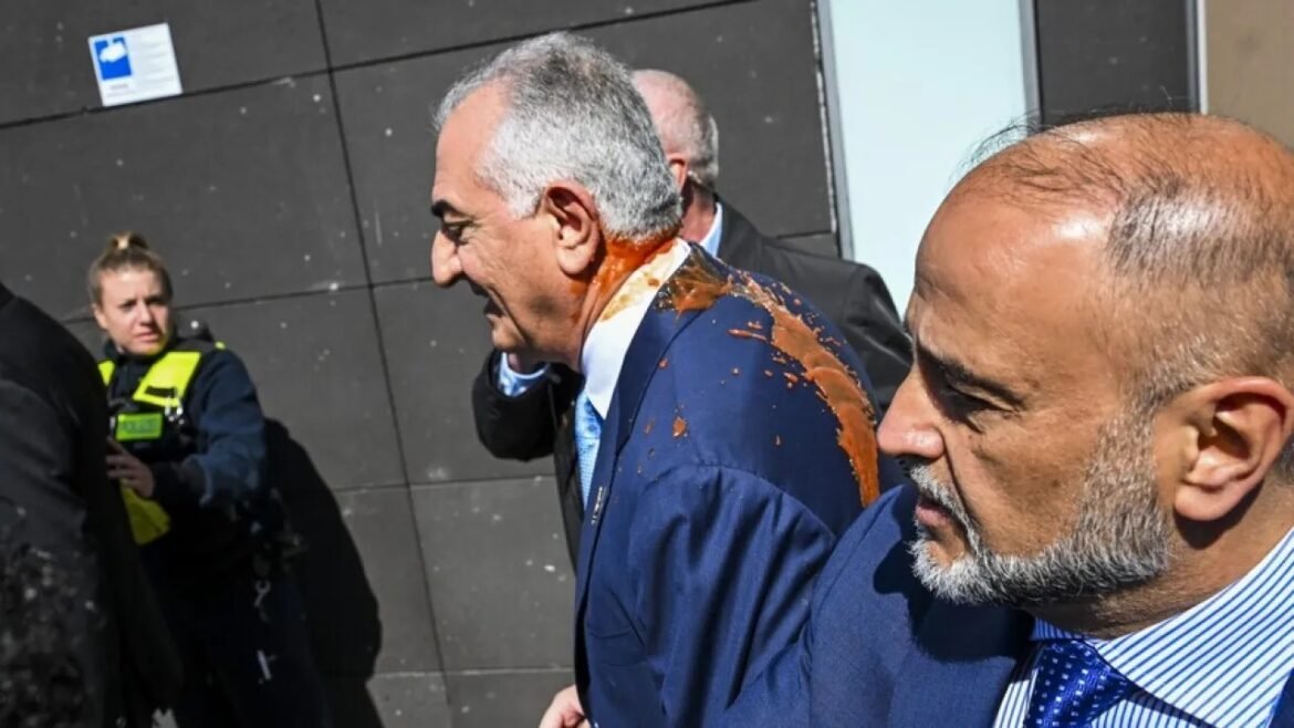 Two men in suits walk outdoors; one man has brown liquid splattered on his shoulder while security stands nearby.