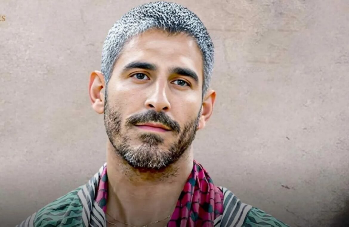 Close-up portrait of a man with salt-and-pepper hair and beard, wearing a patterned scarf, looking at the camera.