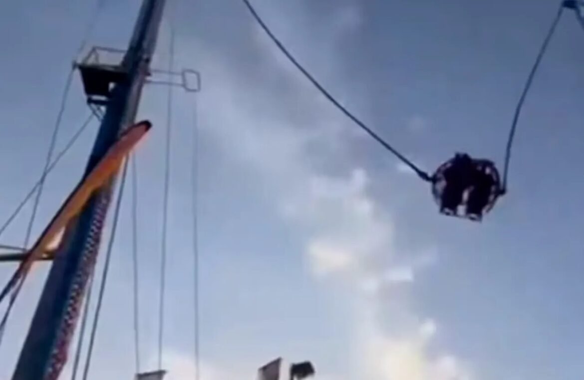 Pendulum-style amusement ride: a circular cage swings high with riders inside as it trails from a tall tower against a blue sky, showing motion and height.