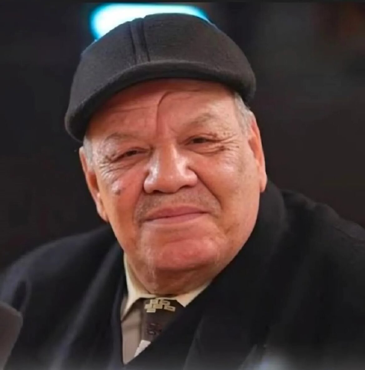 Smiling older man wearing a dark cap and blazer, facing the camera.