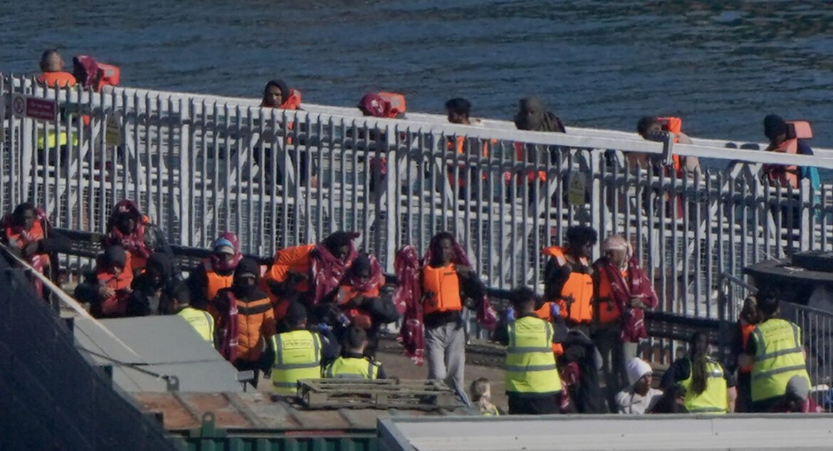 Group of people in bright orange life jackets gathered on a dock by the water, some wearing blankets, with officials in yellow vests nearby.