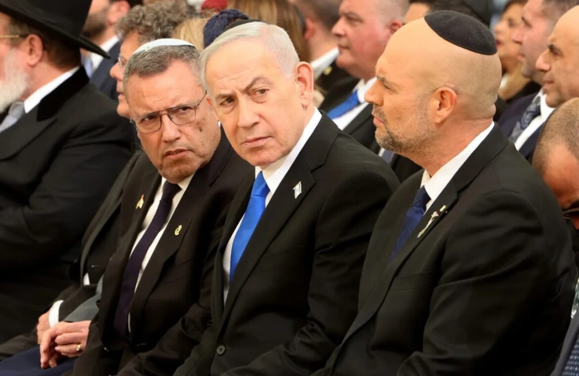Three men in dark suits sit in a formal ceremony, two wearing kippahs, with a crowd behind them and attentive expressions.