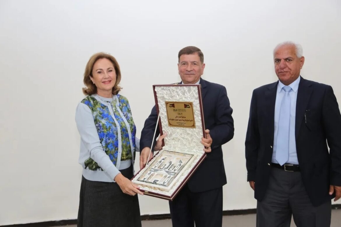 Three adults in formal attire stand in a plain room while presenting and receiving a decorative award box.