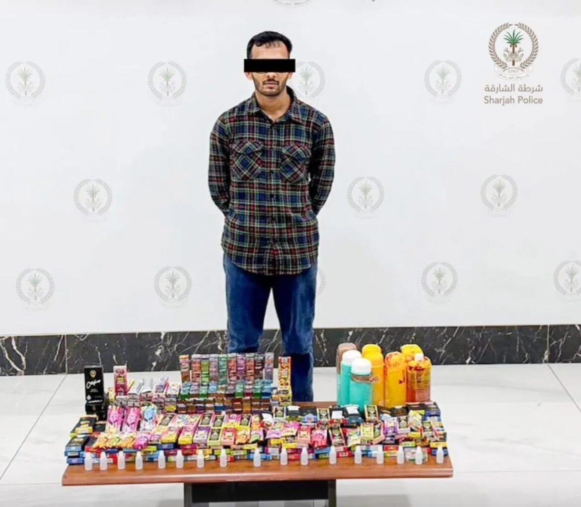 Man with a black bar over his eyes stands behind a table loaded with assorted seized goods in a police backdrop.
